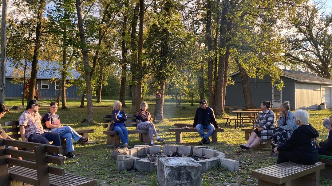 a group of about 10 people are scattered on wooden benches around a stone fire pit. sunlight comes through the tree branches behind them.