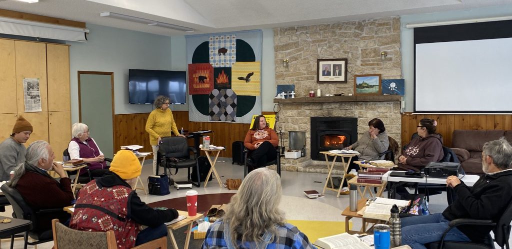 Students sit in a circle of chair in the Sandy-Saulteaux hall. There is a fireplace and a large quilted wall hanging in the background.