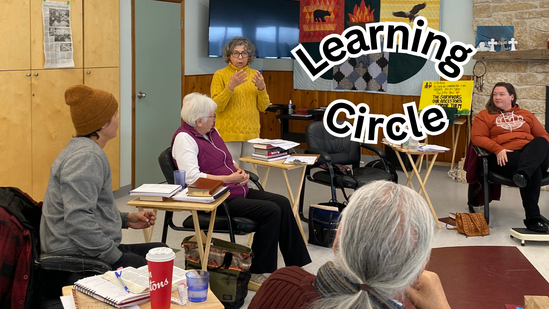Students of varying ages, dressed casually, are seated in chairs, facing in to a circle. Each chair is separated with a small table where papers, books and a Tim's coffee cup are resting. One person is standing and speaking. In the background there is a blank TV screen, a quilted wall hanging and the edge of a stone fireplace.