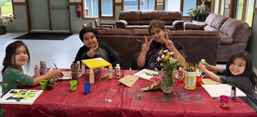 4 children sit around a table with paint supplies, paper, brushes and a vase of flowers. They have big smiles.
